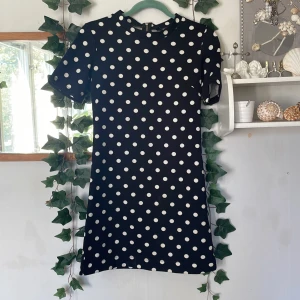 Svart prickig kortklänning från H&M - Säljer en svart kortklänning från H&M Divided med vita prickar. Klänningen har en rak passform, korta ärmar och rund hals. Dragkedja finns i ryggen för enkel påklädning. Perfekt för dig som gillar retrovibbar och lekfull stil.