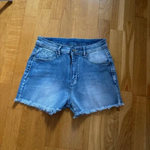 Blå jeansshorts med broderade fickor - Snygga blå jeansshorts med fransad kant och klassisk femficksdesign. Baksidan har broderade detaljer med blommor på fickorna som ger extra stilpoäng. Perfekta för varma dagar och chill häng. Midwaist passform och mjukt denimtyg. Storlek M