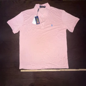 Rosa Polo Ralph Lauren t-shirt - Snygg ljusrosa t-shirt från Polo Ralph Lauren med klassisk pikékrage och tre knappar framtill. T-shirten har korta ärmar och är dekorerad med det ikoniska blå broderade logot på bröstet. Tillverkad i mjuk bomull för skön känsla.
