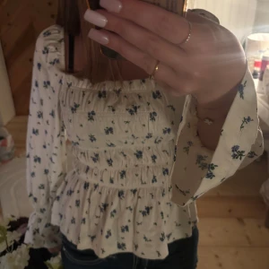 Blommig vit blus med puffärmar - Supersöt blus i vit med blått blommönster och smockad midja. Blusen har fyrkantig halsringning, långa puffärmar och volang nedtill. Materialet känns som mjuk bomull.
