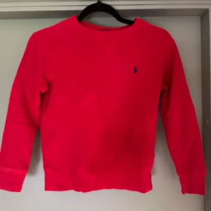 Röd sweatshirt från Polo Ralph Lauren - Snygg röd sweatshirt från Polo Ralph Lauren i storlek S. Tröjan har rund hals, långa ärmar och en liten blå logga broderad på bröstet. Tillverkad i mjuk bomull som är skön mot huden. Perfekt för dig som gillar klassisk och stilren streetwear.