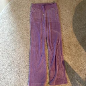 Lila Juicy Couture XS - Säljer ett par lila mjukisbyxor från Juicy Couture i storlek XS. Byxorna har bred resår med snörning i midjan, fickor fram och bak med snygga detaljer och JC-broderi. Materialet är mjukt och skönt, perfekt för chill dagar. Färgen är ljuslila och byxorna har en loose passform.