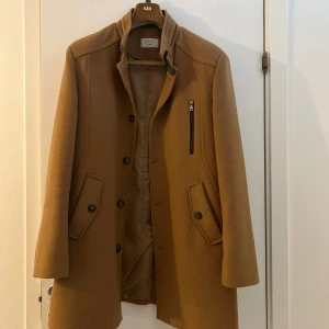 Brun kappa från Aquard - Snygg brun kappa från Aquard med klassisk krage och knappstängning. Jackan har en rak passform, långärmade ärmar och praktiska fickor med lock samt en cool bröstficka med dragkedja. Perfekt för dig som gillar stilrena och tidlösa plagg.