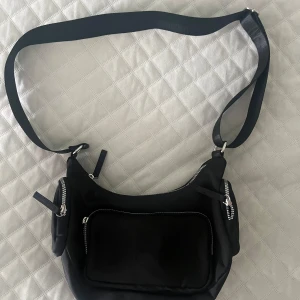 Svart axelväska med dragkedjor - Snygg svart axelväska med bred  justerbar rem och flera praktiska fack med silverfärgade dragkedjor. Väskan har en mjuk, rundad form och ett stort ytterfack framtill. Perfekt för att bära med sig allt nödvändigt och passar till många olika stilar.