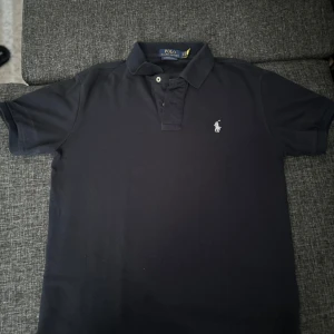 Svart Ralph Lauren Polo - Svart t-shirt från Polo Ralph Lauren med klassisk pikékrage och två knappar framtill. Slim fit-modell med vit broderad logga på bröstet. Tillverkad i mjuk bomull som känns skön mot huden. Perfekt för dig som gillar stilren och enkel design.