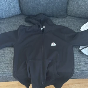Svart Moncler hoodie med dragkedja - Svart hoodie från Moncler med klassisk logga på bröstet. Tröjan har huva, hel dragkedja och stora fickor framtill. Materialet känns mjukt och är troligtvis bomull eller polyester. Perfekt för chill dagar och streetwear-stil.
