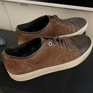 Bruna Lanvins med glans  - Snygga bruna sneakers från Lanvin Paris med mörkbrunt tåparti och vit platt sula. Skorna har snörning och är tillverkade i skinn med en lyxig känsla. Perfekt för dig som gillar stilrena och exklusiva sneakers med en unik touch.