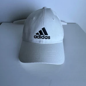 Adidas keps - Snygg vit keps från Adidas med klassisk svart logga framtill. Kepsen har böjd skärm, ventilerande hål och justerbar passform. Tillverkad i bomull för en skön känsla och stilren look. Perfekt för dig som gillar sportig och enkel design.