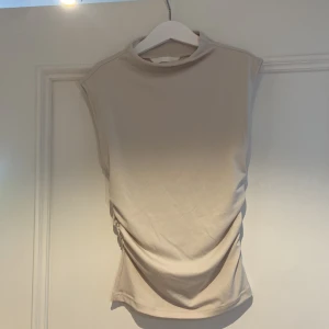 Beige omlott topp från H&M XS - Snygg beige topp från H&M i storlek XS. Modellen är ärmlös med hög hals och har en cool omlott-detalj med snörning på sidan som ger en draperad effekt. Materialet är mjukt och stretchigt, perfekt för att skapa en clean och trendig look.