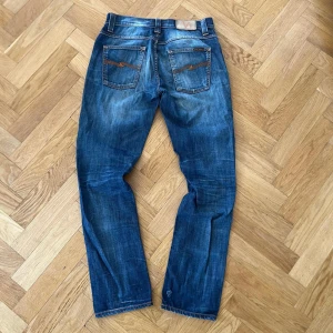Nudie jeans - Snygga blå jeans från Nudie Jeans med klassisk femficksdesign och coola slitningar. Jeansen har orange sömmar och Nudie-logga på bakfickan. Passformen är rak och normal, perfekt för dig som gillar en avslappnad stil. Materialet är slitstark denim i bomull.