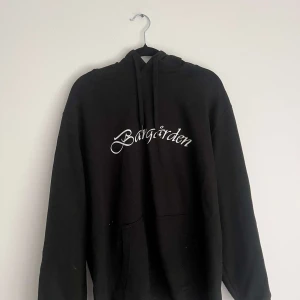 Svart Bangården hoodie med tryck - Svart hoodie från Bangården Customs med vit text på bröstet och stort krontryck på ryggen. Tröjan har huva med snörning och en klassisk känguruficka framtill. Perfekt för dig som gillar streetwear och vill sticka ut med coolt motiv. Storlek 2xl men sitter som medium eller small.