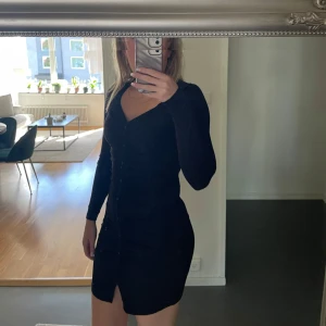 Svart v-ringad kortklänning - Säljer en svart, figurnära kortklänning med v-ringning och långa ärmar. Klänningen har knappar framtill och en liten slits nedtill som ger en snygg detalj. Perfekt för dig som gillar stilrena och enkla plagg med en twist.