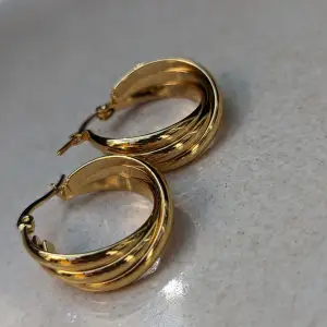 Säljer ett par unika och moderna örhängen i rostfritt stål. Dessa örhängen har en trendig hip hop-design med en dinglande ring och en trippel ring-design som ger en hög 3D-textur. De är guldfärgade och utstrålar elegans och stil  Rostfritt stål,ny