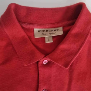 Röd piké från Burberry  - Pikétröja från Burberry. Tröjan är i fint skick men har ett litet märke efter ett strykjärn där tyget har blivit lite glansigare 