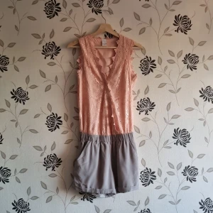 Vila playsuit, Strl: S, sommar, fest, v-ringad, spets, rosa, jumper, byxdress, - Jättesöt och somrig rosa och grå playsuit från Vila, storlek small. Rosa spets och grått glansigare tyg, resår i midjan och man kan ha ett bälte i den. Dold knäppning i fram. Nyskick då den ej blivit använd! OBS! Jag kan inte skicka något vecka 26.