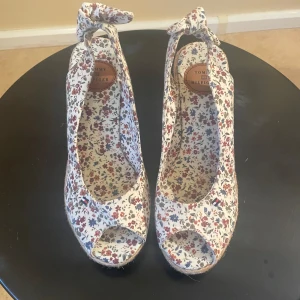 Blommiga kilklackar från Tommy Hilfiger - Ett par blommiga kilklackar från Tommy Hilfiger med en knytning vid ankeln. Skorna har en kilklack täckt med ett rep-liknande material och en öppen tå-design. Färgerna på blommönstret inkluderar röd, blå och beige.