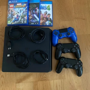 PlayStation 4 med tre handkontroller och tre spel - En PlayStation 4-konsol med tre handkontroller (två svarta och en blå) samt tre spel: LEGO Marvel Super Heroes 2, Star Wars Battlefront II och The Sims 4. Konsolen kommer med nödvändiga kablar och en strömadapter.