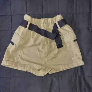 Ett par beige shorts med svarta detaljer och fickor. Shortsen har en elastisk midja och svarta band som dekoration.