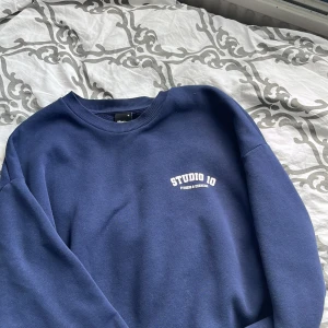 Blå sweatshirt från Studio 10 - En bekväm blå sweatshirt från Studio 10 med texten 'STUDIO 10 FITNESS & GYM' tryckt på bröstet. Tröjan har långa ärmar och ribbstickade muddar vid ärmslut och nederkant.