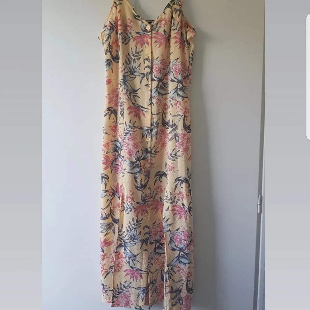 En pastell gul och lång sundress med blommigt motiv i korall färg och svartvitt. Framsidan har knappar hela vägen ner och sen två slit på var en sida för att visa lite extra ben.. Klänningar.