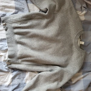 Grå tröja med dragkedjor - En grå tröja med rund hals och dragkedjor på sidorna. Tröjan har långa ärmar och ribbstickade muddar vid ärmslut och nederkant.