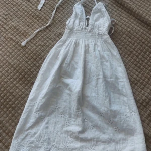 Vit sommarklänning med spetsdetaljer - En vacker vit sommarklänning med spetsdetaljer och broderier. Klänningen har en omlott-design med knytband och en elastisk midja för en bekväm passform. Den är ärmlös och har en öppen rygg.