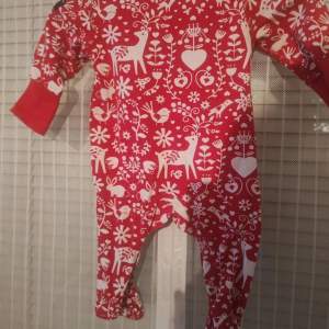 En söt och bekväm röd babypyjamas från Polarn O. Pyret med ett vitt mönster av hjärtan, blommor och renar. pyjamasen har långa ärmar och muddar vid ärmslut och heltäckande fot