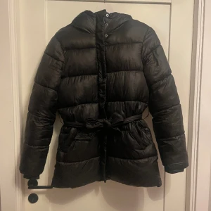 Vinterjacka  - Vinterjacka från Pieces med bälte.  Den har ett hål i högra fickan men är annars i bra skick!  Obs jackan har inget mönster utan den är hel svart med lite glansigt material. Fick inte till en bra bild 