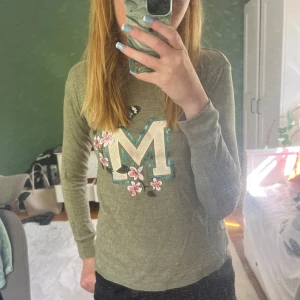 Grön långärmad tröja - Grön tröja med ett M i mitten och en luva. Den har nått tecken på användning och M:et är färgat lite lila därav priset 💜om det är nån fråga om pris eller något annat skriv till mig💖