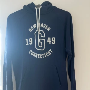 Mörkblå hoodie med tryck - En mörkblå hoodie med vit text och tryck på framsidan som säger 'New Haven Connecticut 1949'. Hoodien har en justerbar huva med vita snören och en magficka. Den är långärmad och tillverkad i ett mjukt material.