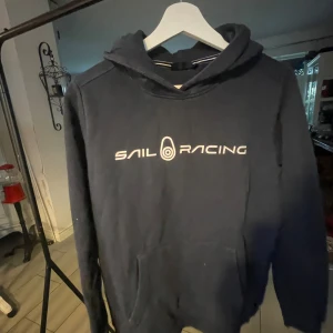 Mörkblå hoodie från Sail Racing - En mörkblå hoodie från Sail Racing med vit text på bröstet. Hoodien har en stor ficka framtill och en justerbar huva.