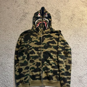 Bape Hoodie - Bape 1st edition hoodie Köpt i Miami i retail butik! Storlek L, sitter något mindre som bape brukar. Skick: 7,5/10, inga fläckar eller crackings med väl använd. Extremt limiterade exemplar samt 1st edition på camo-shark släppen.    