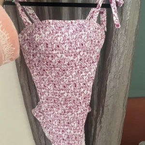 Baddräkt/ body - Urgullig baddräkt, fungerar även som body! Vit med vinröd/lila detaljer. Rosetter på axlarna som man knyter själv 🎀 Smockad så passar nog XS-M 💗  Ilägg som går att ta ut om man önskar