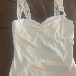 Vit topp med spetsdetaljer - Säljer en superfin vit topp med spetsdetaljer på axelbanden. Toppen har en snygg rynkning vid bysten och är perfekt för sommarens alla fester. Den är i nyskick och passar perfekt till både vardag och fest. Materialet känns mjukt och skönt mot hude