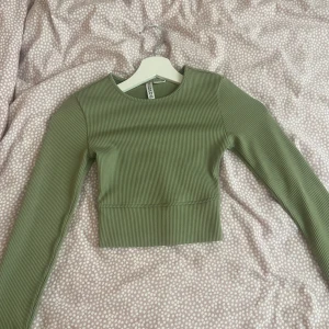 Grön ribbad topp från Divided - Säljer en snygg grön ribbad topp från Divided. Den är i storlek XXS och har långa ärmar. Toppen är croppad och har en tight passform. Materialet är mjukt och stretchigt, vilket gör den bekväm att bära. Är skön att både träna i och att ha vardagligen 