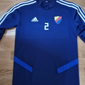 Blå långärmad fotbollströja från Adidas - Säljer en blå långärmad fotbollströja från Adidas med vita ränder på ärmarna och ett klubbmärke på bröstet. Tröjan har nummer 2 tryckt på framsidan. Perfekt för fotboll med vänner eller nåt annat.