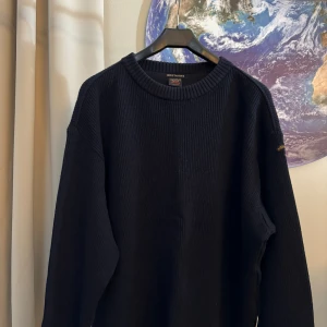 Svart stickad tröja från Paul & Shark - Säljer en snygg och bekväm svart stickad tröja från Paul & Shark. Tröjan har en klassisk design med ribbade detaljer på ärmarna och runt halsen. Perfekt för höst och vinter, den håller dig varm och stilren. Märket Paul & Shark är broderat på ärmen för en extra touch av lyx. Passar bra till både vardag och fest.