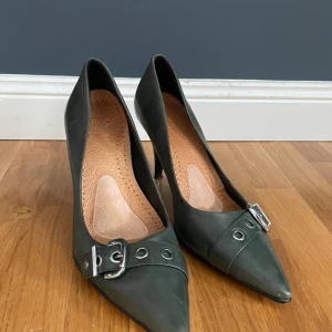 Mörkgröna klackar med buckeldetalj  - Ett par mellan-höga klackar med fint grönt äkta läder och en silvrig detalj. Från 5th Avenue