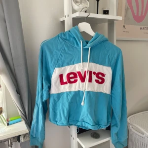 Blå hoodie från Levi's - En blå hoodie från Levi's med en vit och röd logotyp på framsidan. Hoodien har en justerbar dragsko vid huvan och långa ärmar.