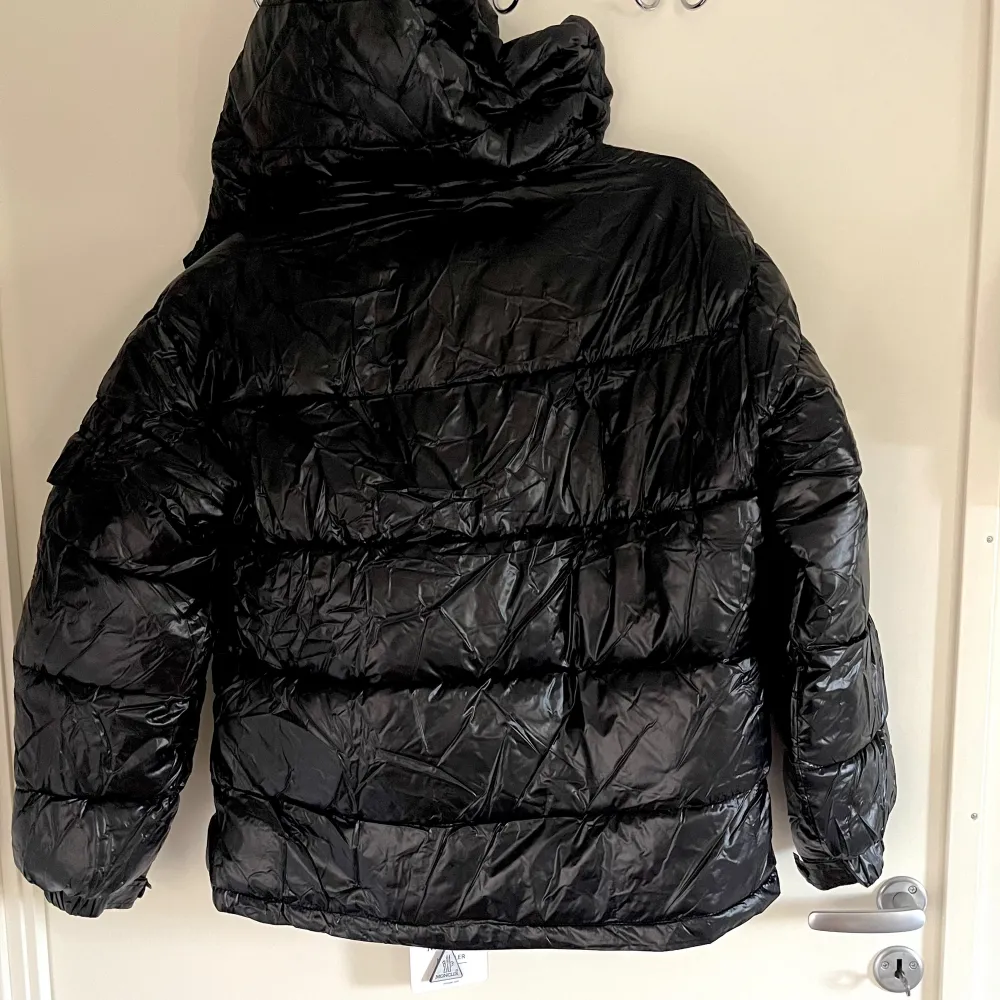 En svart dunjacka från Moncler med huva och dragkedja. Jackan har flera fickor med dragkedjor och en logotyp på vänster ärm. Insidan av jackan har en serieteckning.. Jackor.