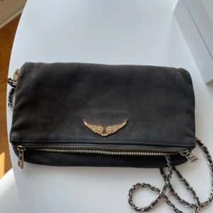 Svart axelväska från Zadig & Voltaire - En elegant svart axelväska från Zadig & Voltaire i mocka. Väskan har en guldfärgad vingdetalj på framsidan och en dragkedja längs botten. Insidan är fodrad och har en innerficka.