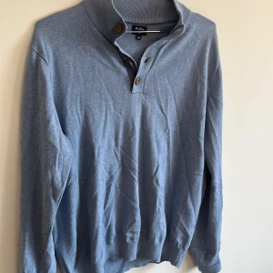 Blå tröja från Mr. Blue - En blå tröja från Mr. Blue med knappar vid halsen. Tröjan har långa ärmar och ribbstickade muddar vid ärmslut och nederkant.