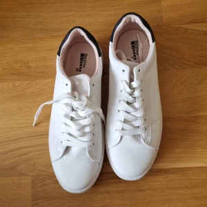 Vita sneakers från Bershka - Ett par vita sneakers från Bershka i storlek 39. Skorna har snörning och en svart detalj på hälen. De är i bra skick och passar perfekt för vardagsbruk.