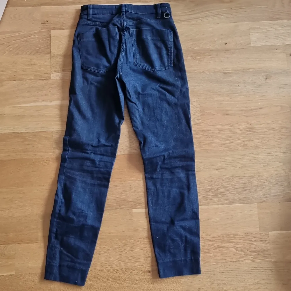 Ett par mörkblå jeansbyxor från Stockh LM i storlek 34. Byxorna är tillverkade av 90% bomull, 6% polyester och 2% elastan, vilket ger dem en bekväm och stretchig passform. De har en klassisk design med fem fickor och knappstängning.. Jeans & Byxor.