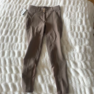 Beige ridbyxor från CRW - Ett par beige ridbyxor från CRW tillverkade av miljövänlig polyester. Byxorna har en hög midja och är designade med knappar och dragkedja framtill. De har även förstärkningar på insidan av benen för extra hållbarhet och komfort vid ridning.