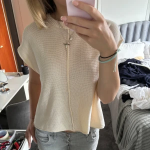 Beige stickad topp - En beige stickad topp med korta ärmar och en dekorativ knäppning framtill. Toppen har en lös passform och är bekväm att bära.
