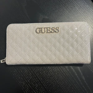 Guess plånbok i nyskick  - Denna praktiska plånbok är perfekt för att hålla kort, kontanter & mynt organiserade. Matchar perfekt med guess väskan jag också säljer. Plånboken har tre fack samt ett myntfack på baksidan. Enbart provad, aldrig använd. 