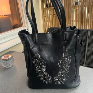 Svart väska med vingar  - Säljer nu min väska som inte kommer till användning något mer. Väskan är i fint skick men använd många gånger. Långt band ingår 🤍