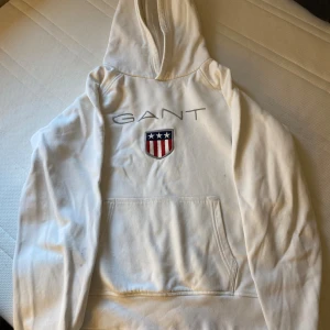 Vit hoodie från GANT - En vit hoodie från GANT med en känguruficka framtill och ett broderat märke på bröstet. Hoodien har en justerbar huva och långa ärmar.