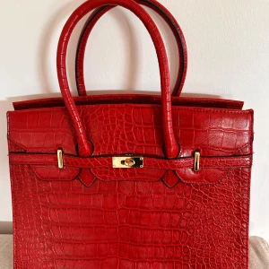 Röd handväska i krokodilmönster - En elegant röd handväska med krokodilmönster. Väskan har två handtag och en guldfärgad låsdetalj på framsidan. Baksidan har en dragkedjeficka. Insidan är rymlig med flera fack för förvaring. Sparsamt använd